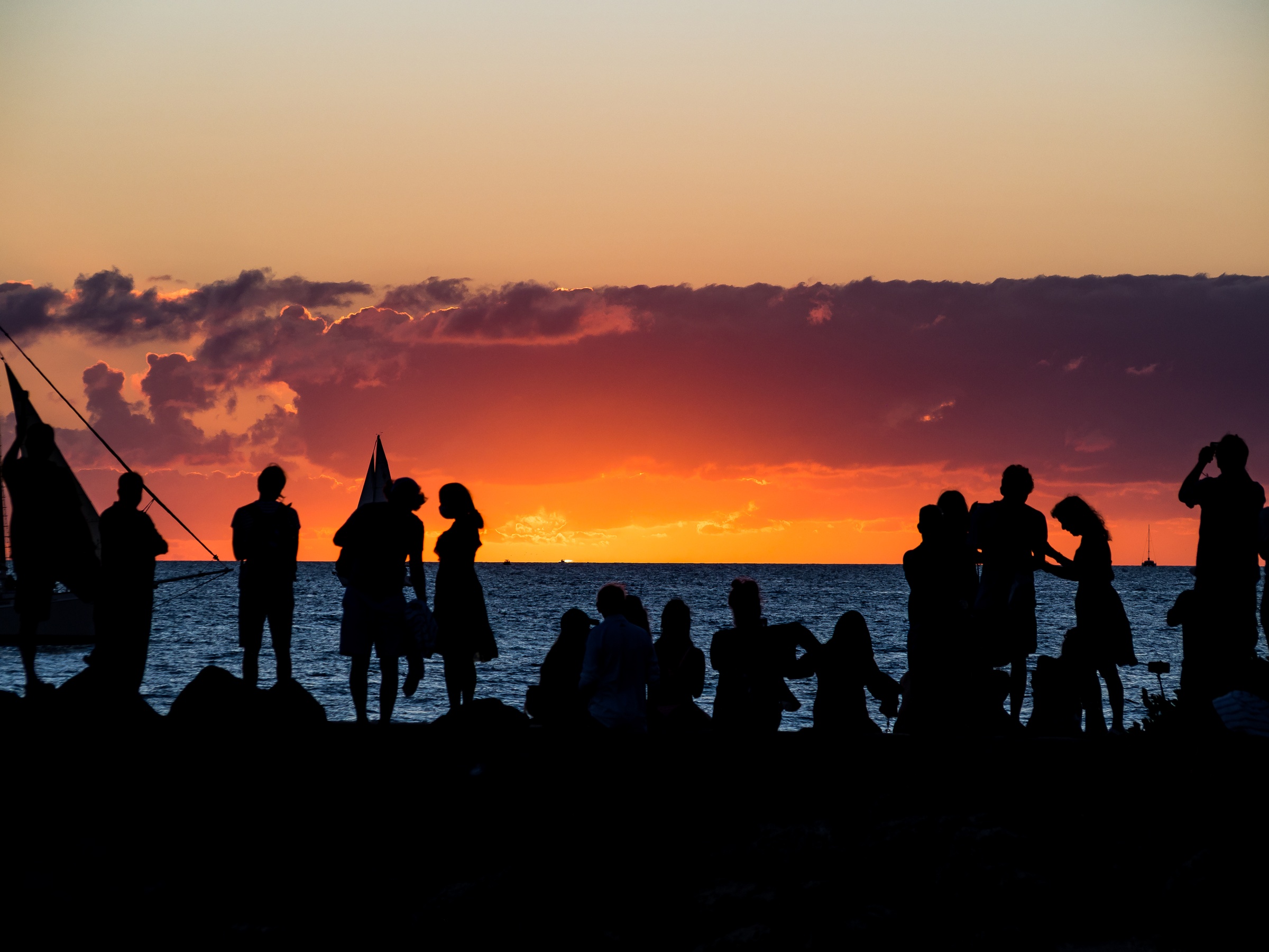 Silhouetted people stand on a shoreline against a colorful sunset sky over water.