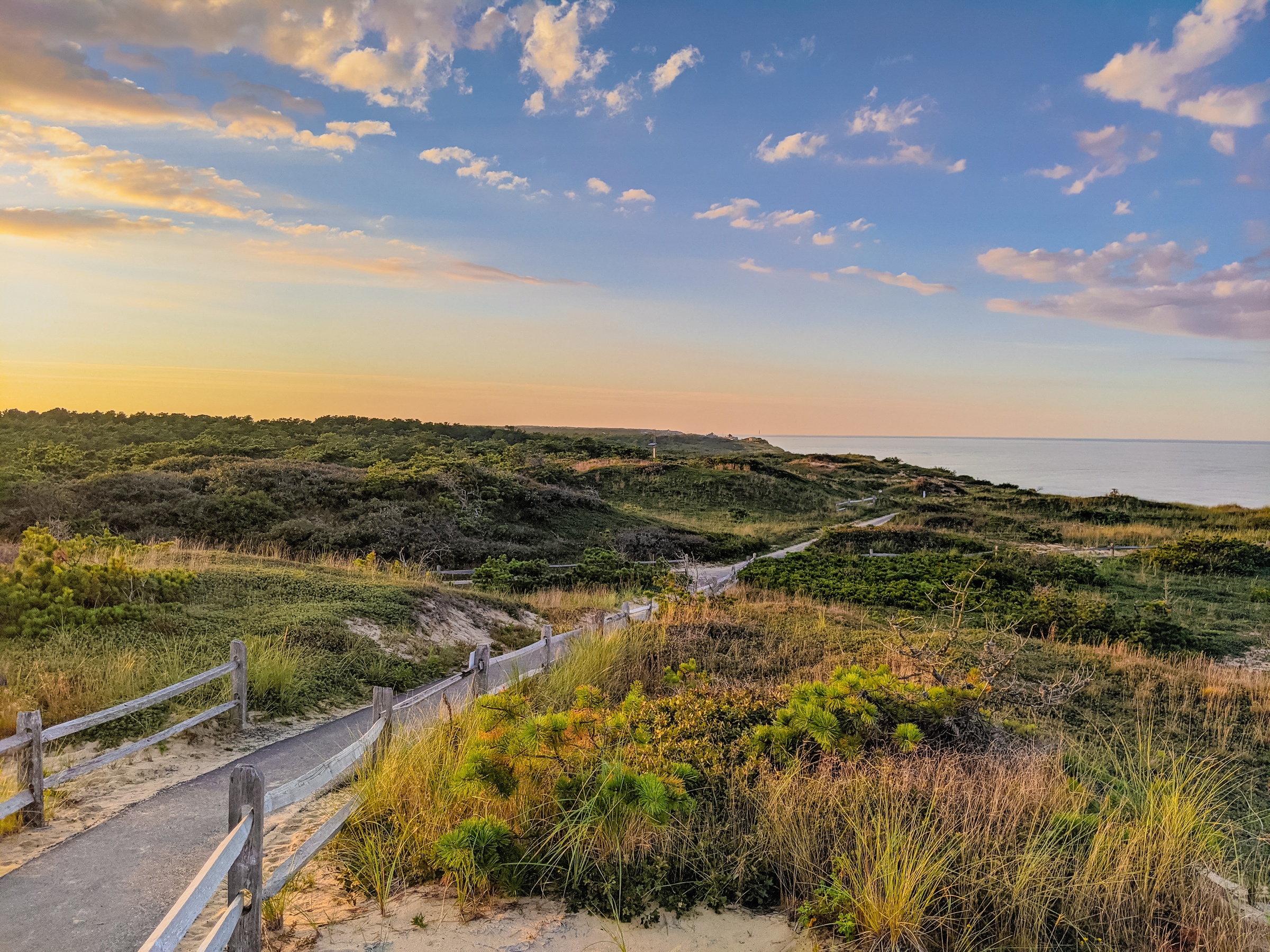 A wooden boardwalk winds through coastal dunes toward the ocean under a colorful sky.