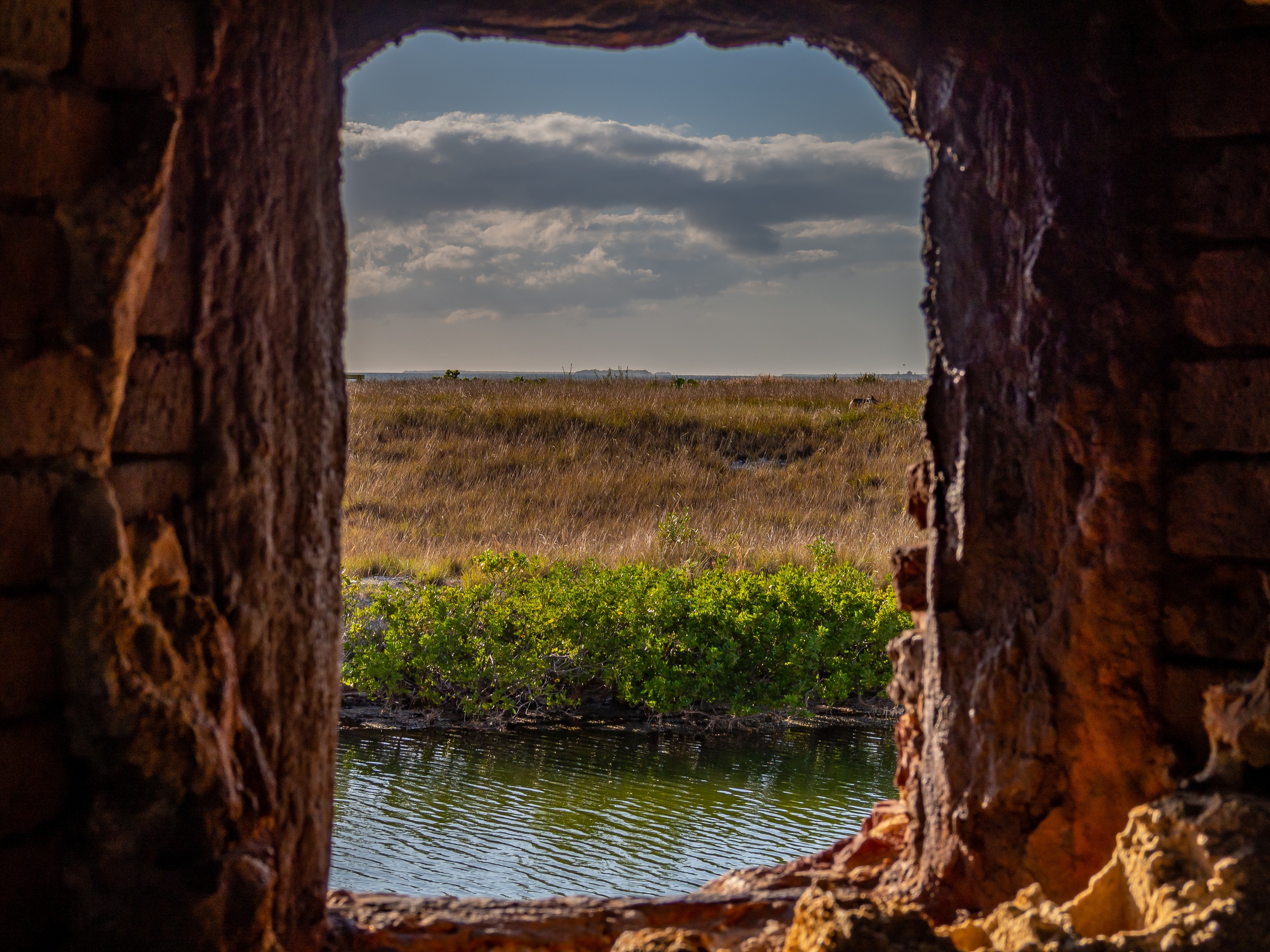 A view of water, greenery, and grasslands through a rough opening in a brick wall.
