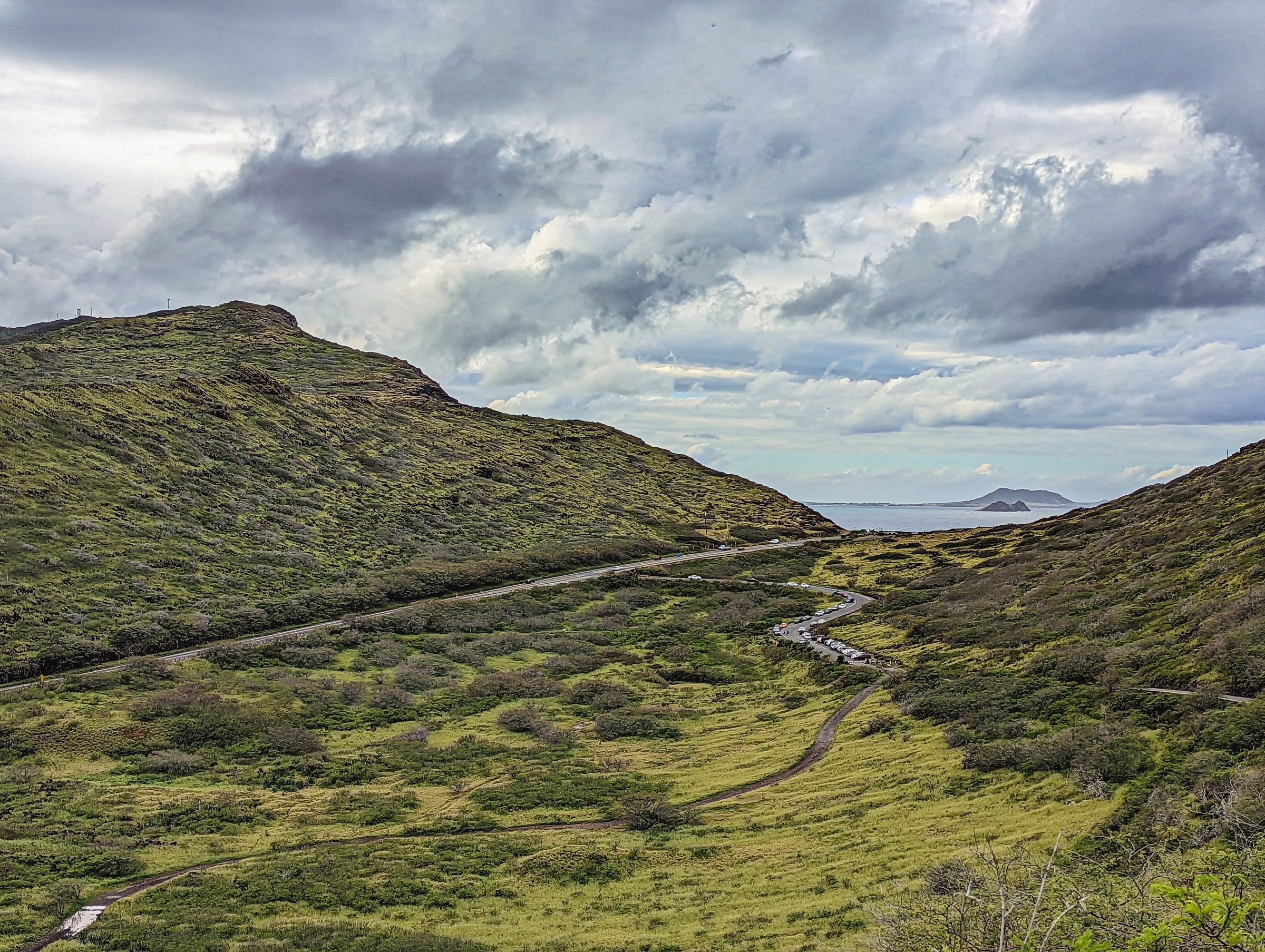 A winding road traverses a green valley between hills under a cloudy sky overlooking the ocean.