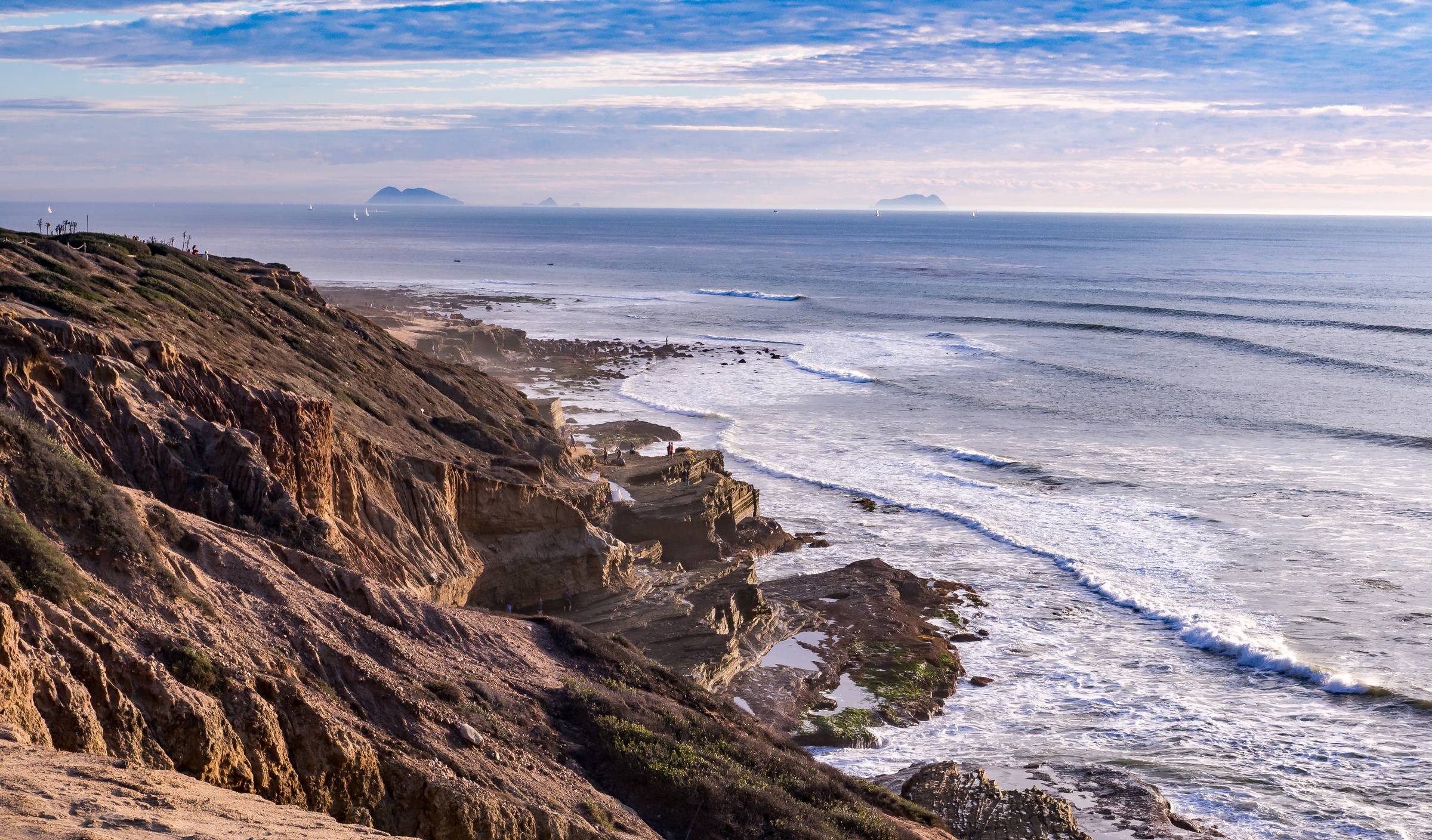 Rugged cliffs overlook an ocean with white waves and distant islands under a cloudy sky.