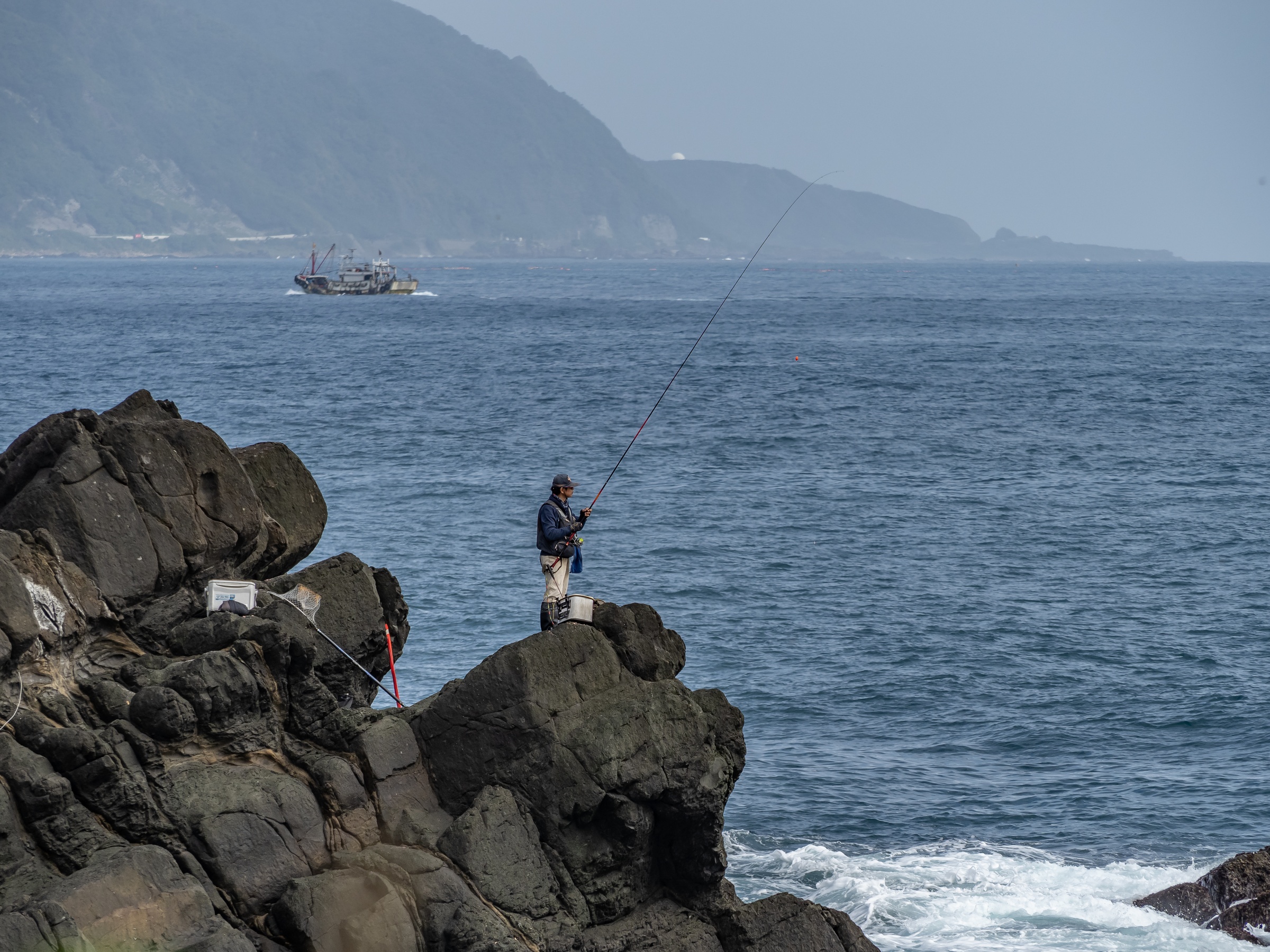 A person fishes from a rocky outcrop by the sea with distant mountains and a boat.