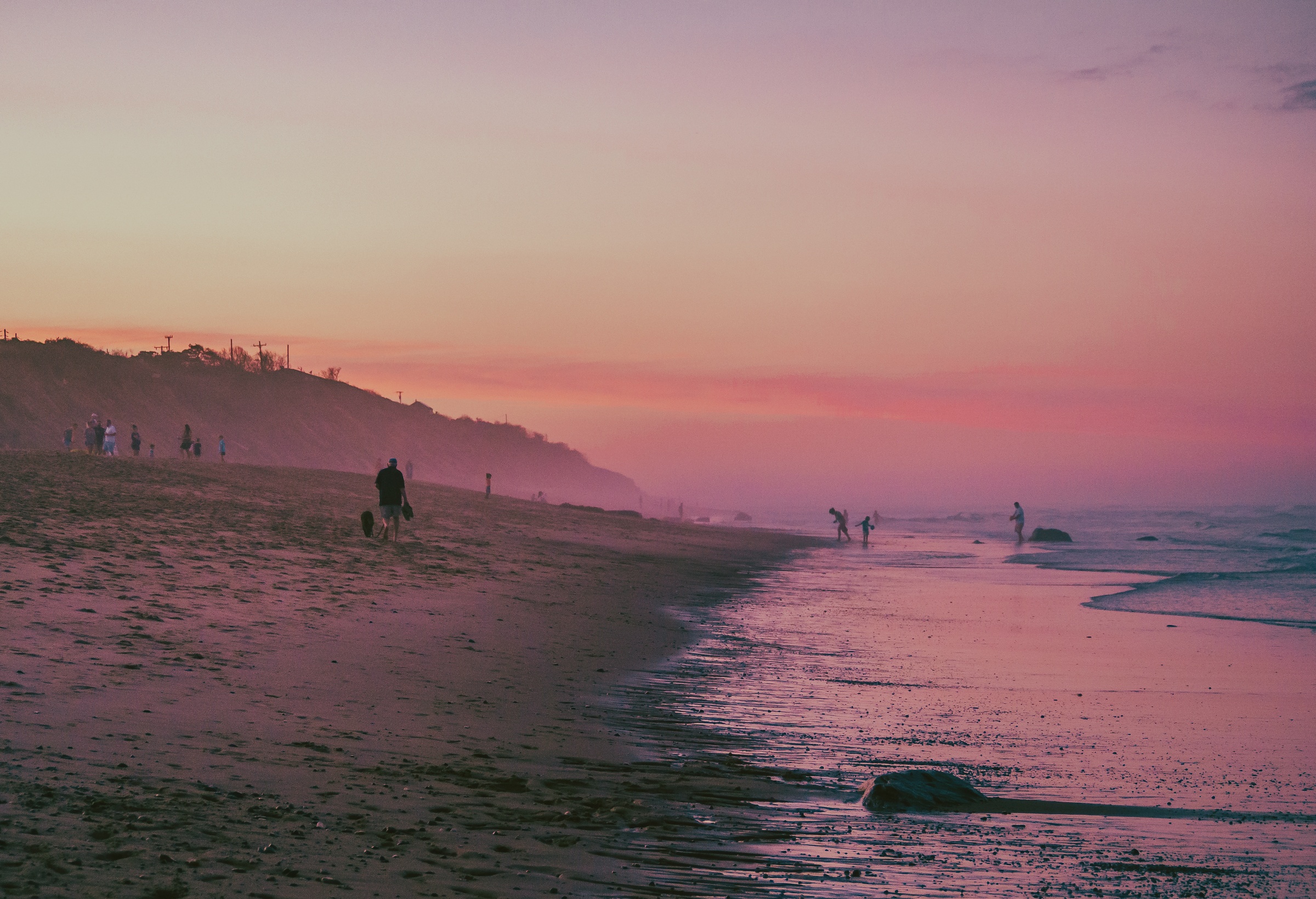 People walk along a sandy beach under a pink and purple sky.