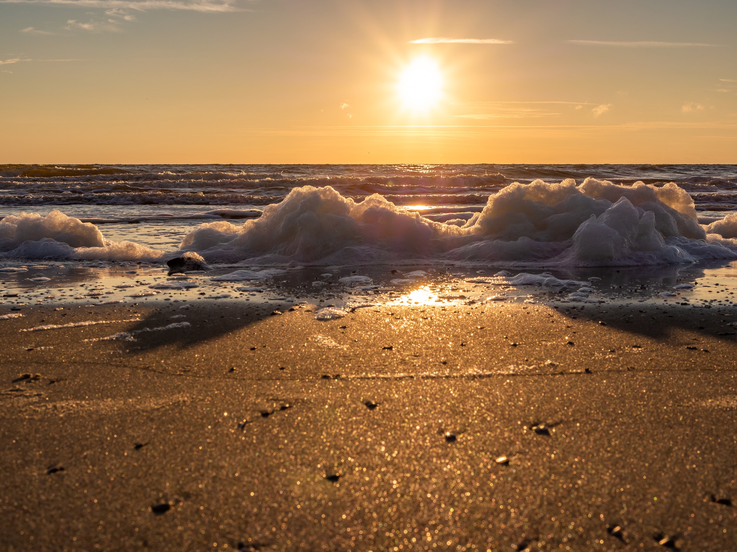 The sun sets over a sandy beach with white sea foam on the shore.