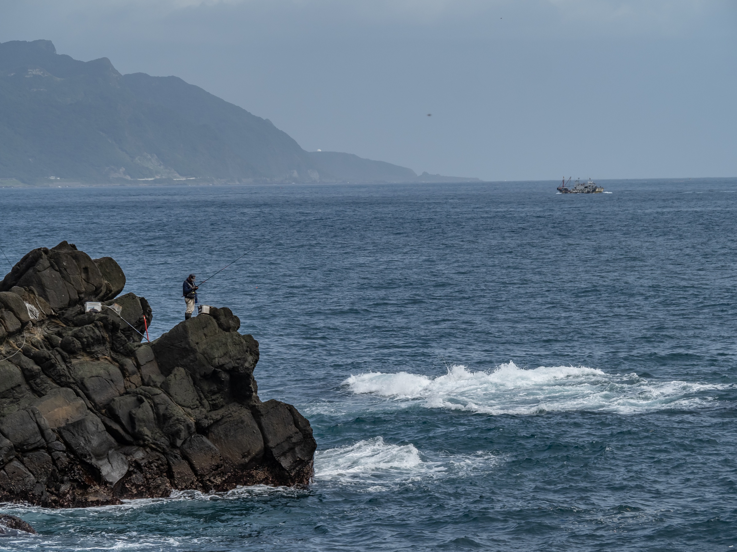 A person fishes from dark rocks by the sea with distant mountains and a small boat.