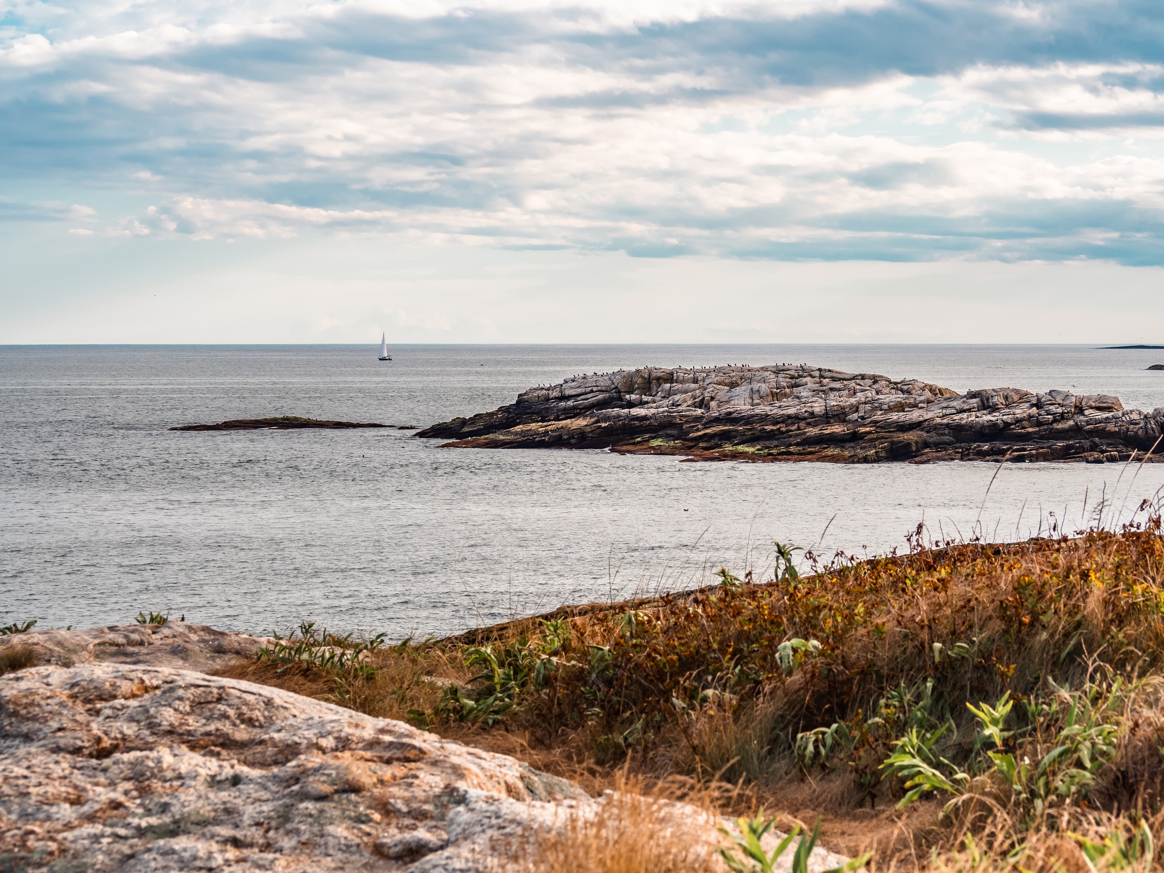 A lone sailboat on the ocean near a rocky coastline under a cloudy sky.