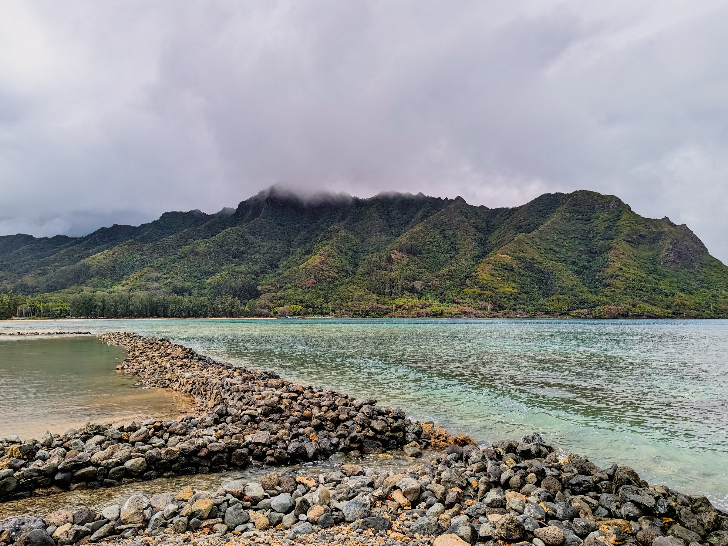 A rocky breakwater extends into turquoise water beneath lush green mountains and a cloudy sky.