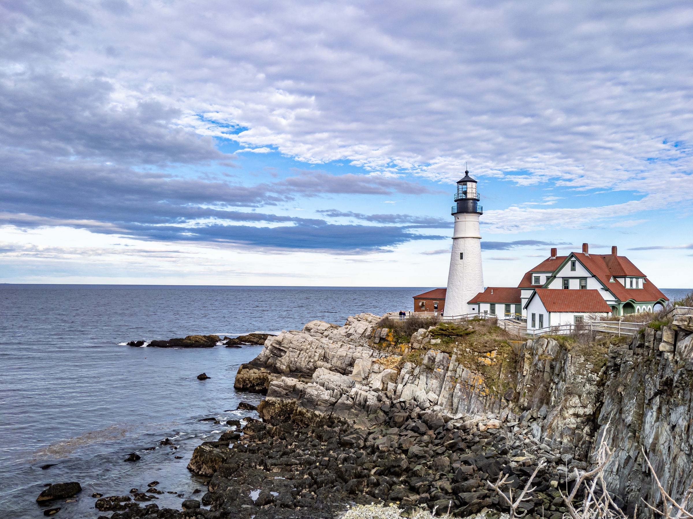 A white lighthouse and red-roofed house stand on a rocky cliff overlooking the ocean.