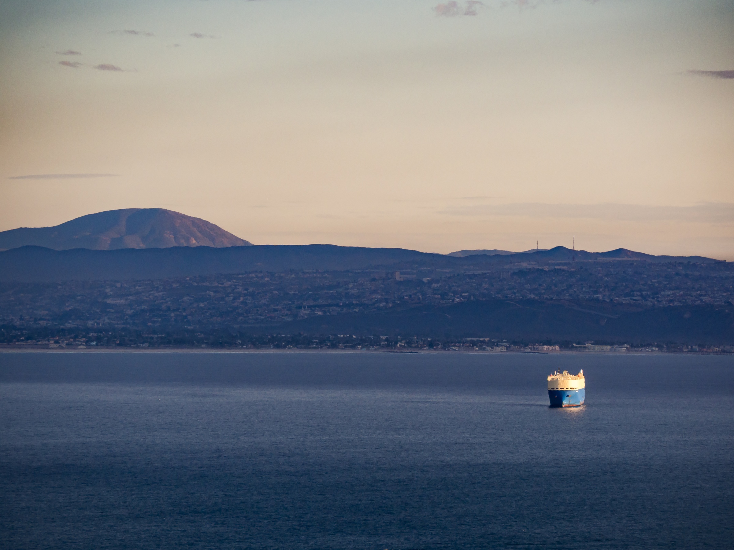 A large ship floats on a calm blue sea before a distant mountainous coastline.