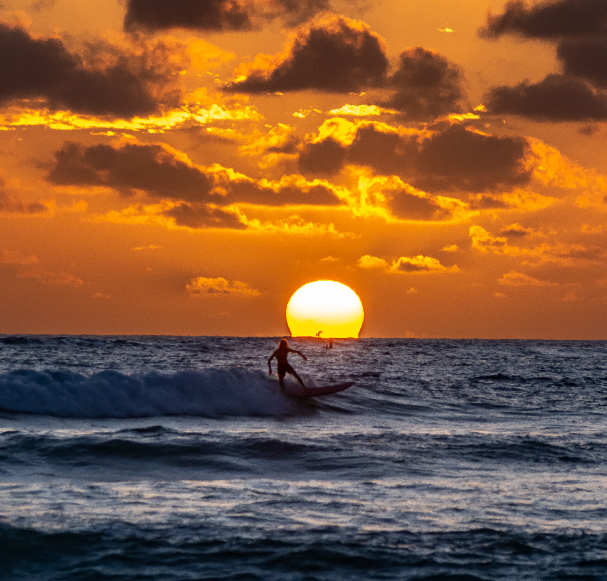 A surfer rides a wave against a large sun and orange cloudy sky.