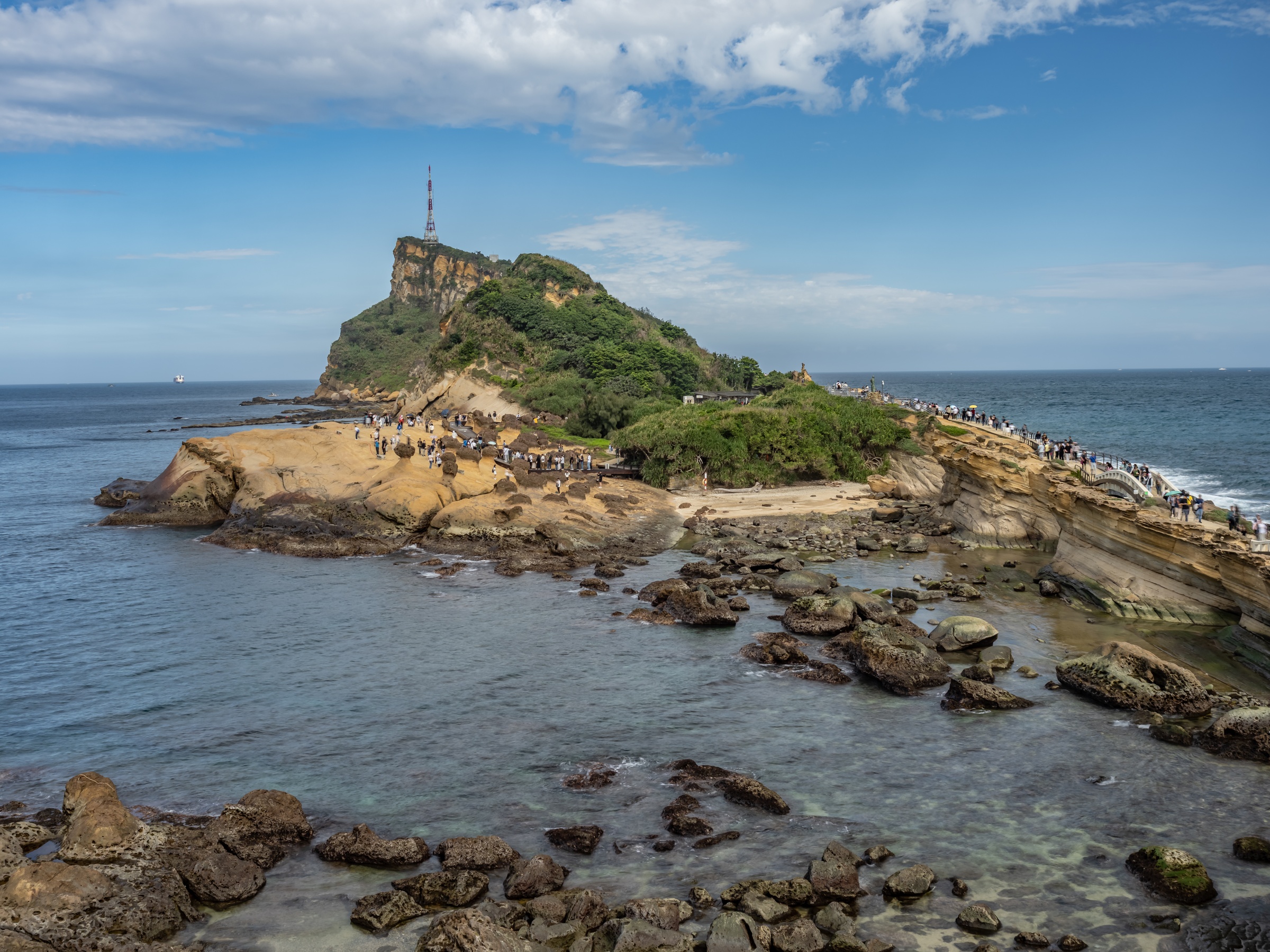People walk along a rocky coastal outcrop topped with a green hill and antenna beside the ocean.