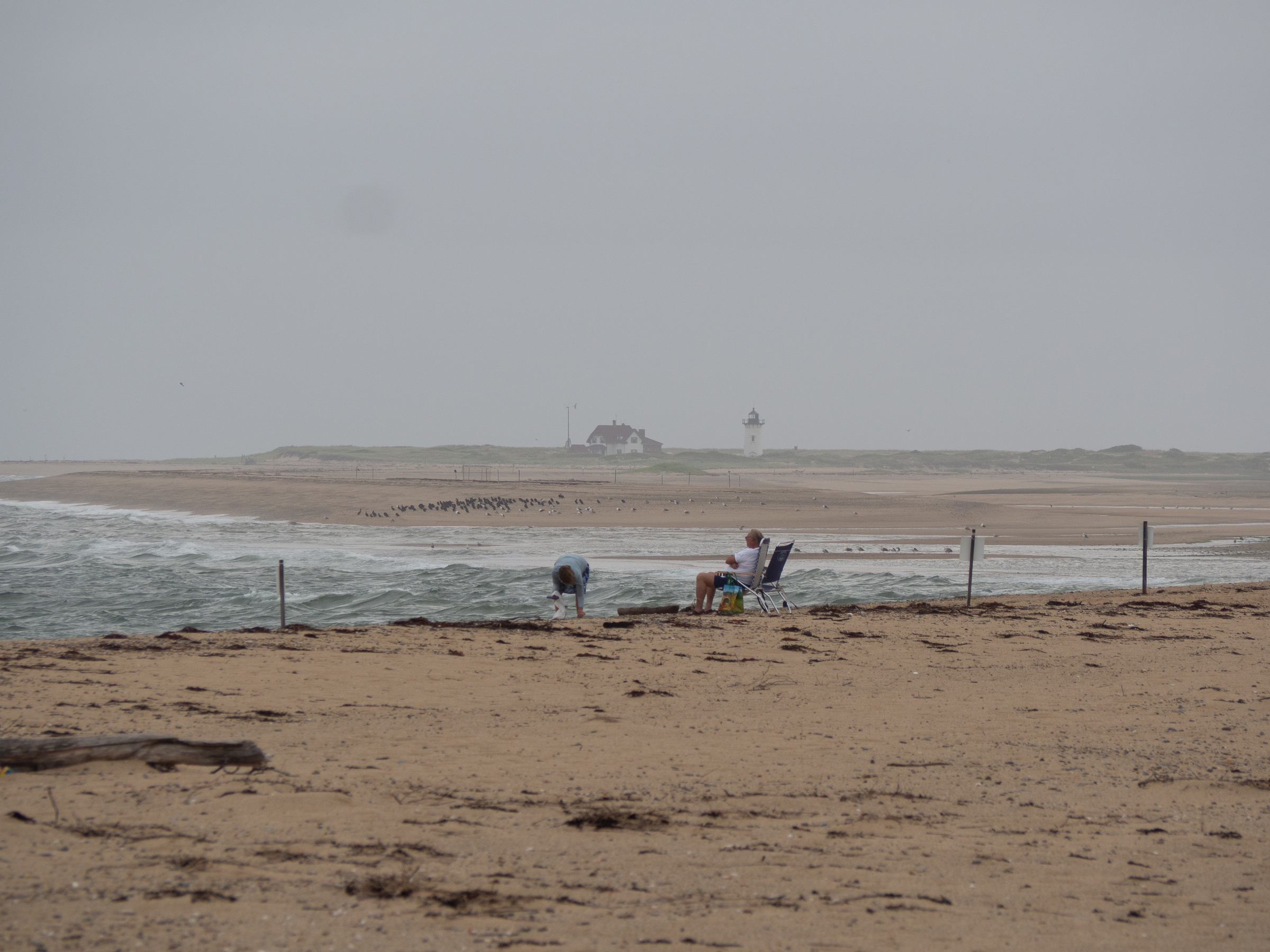 Two people stand and sit on a sandy beach beneath an overcast sky with a distant lighthouse.