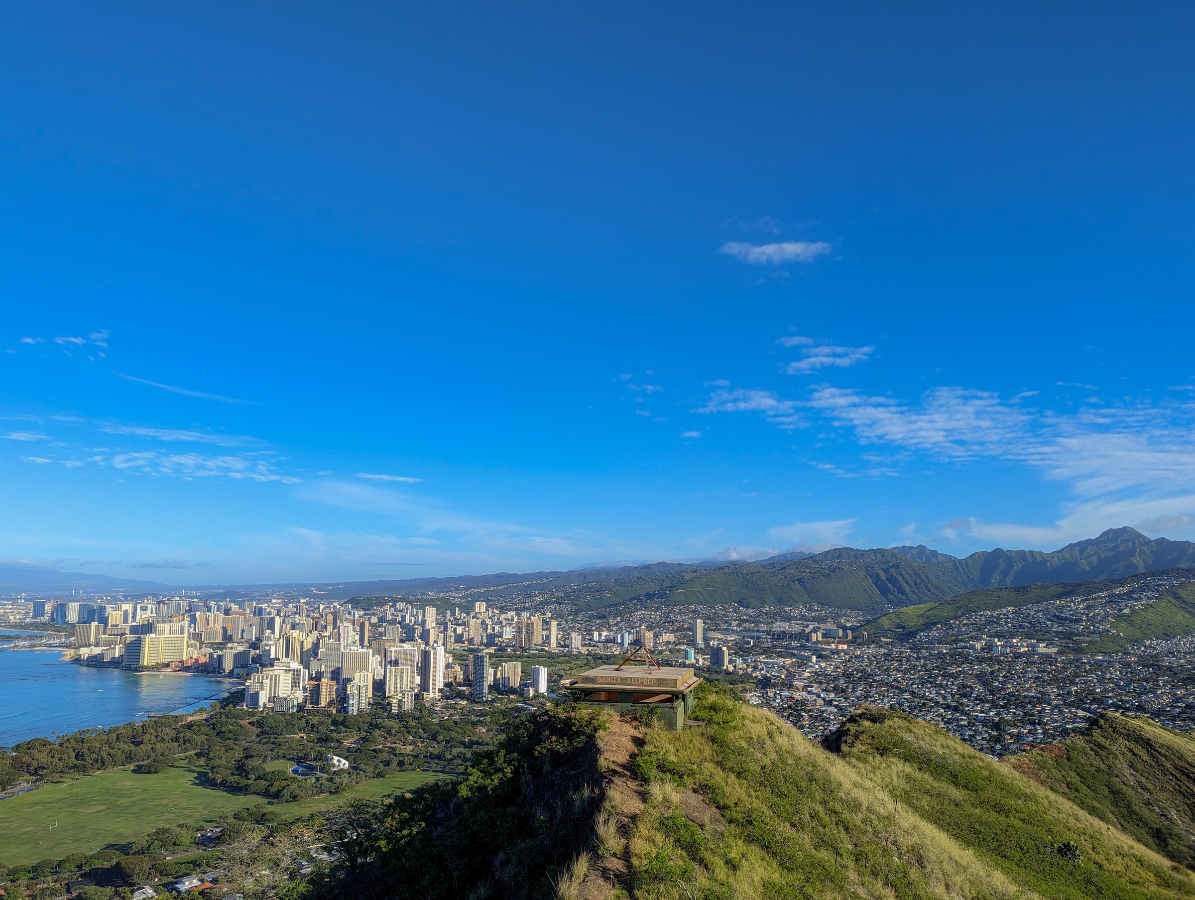 A small structure on a grassy ridge overlooks a sprawling coastal city and distant mountains.
