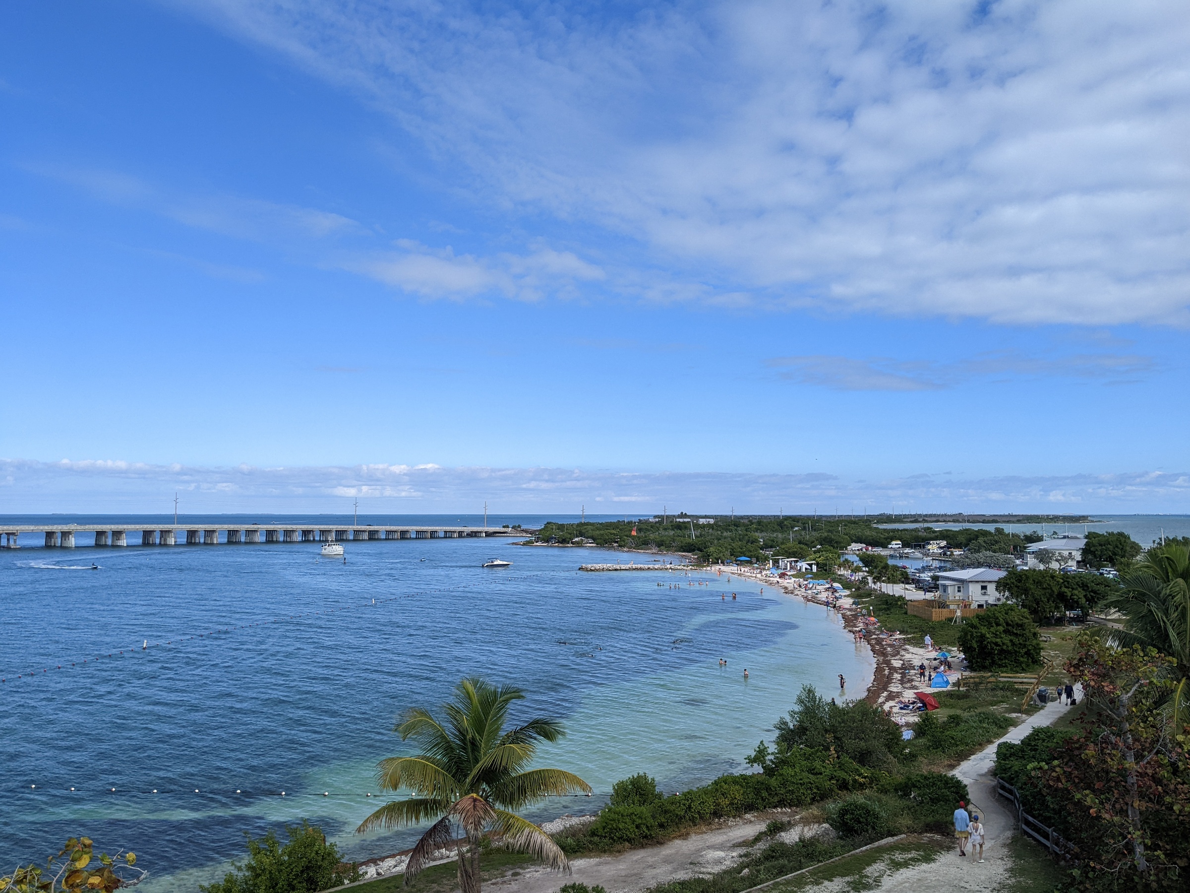 A tropical beach with turquoise water, palm trees, and a long bridge in the distance.